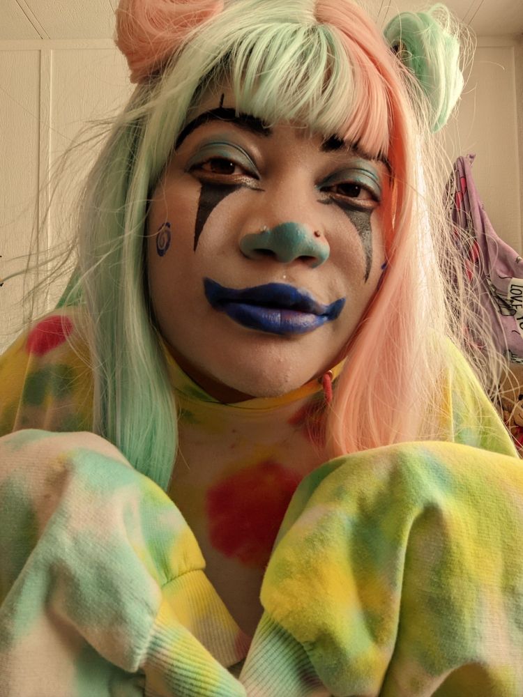 A closeup image of me with my hands held up within hoodie sleeves. My face is covered in white grease paint. My eyebrows are blacked out and have small black lines above them. My eyelids are painted a light teal. There is very light silver sparkly paint beneath my eyes, outlined by a black line. From this line, there are black triangles going down my cheeks. The tip of my nose is painted teal. There is a blue spiral on both of my cheeks. My lips are painted blue and extended into a clown smile. 
My hoodie is a sort of rainbow tie-dye oversized adventure. 
I am wearing a half pink, half blueish-greenish wig, with bangs and space buns in the same half and half colors.