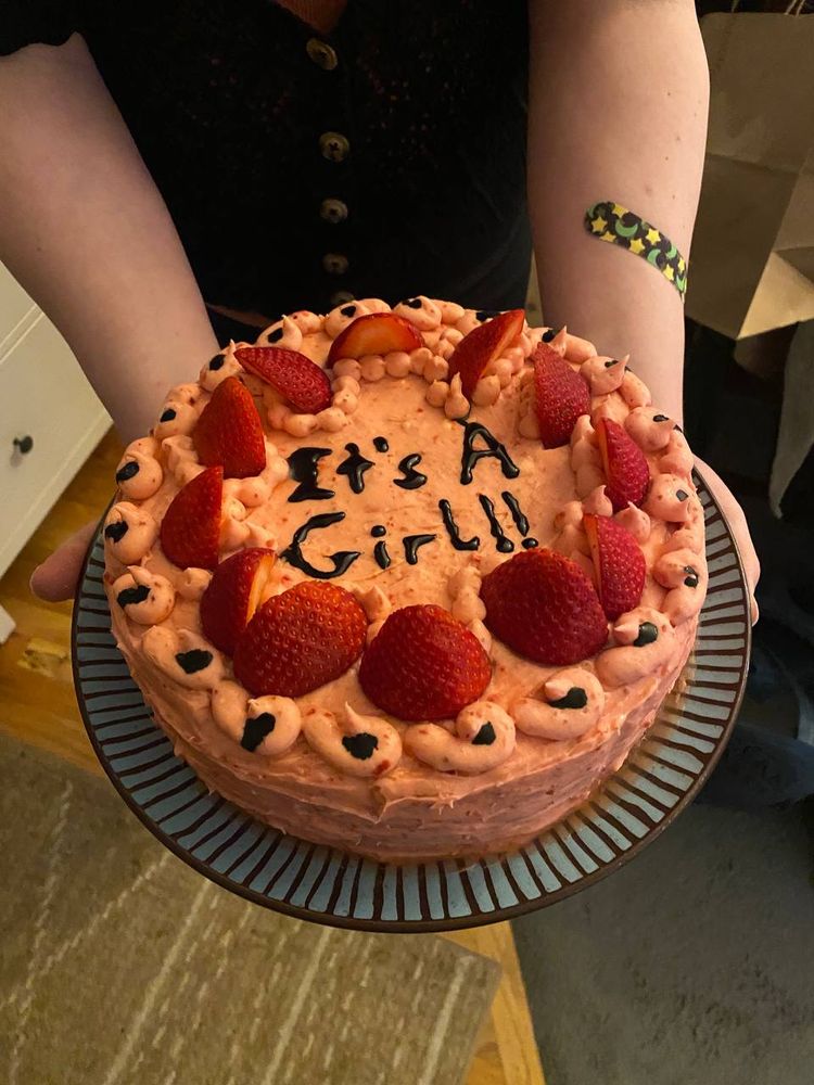 A picture of May's girlfriend holding a cute pink cake covered in strawberries, with piped letters that read 'It's A Girl' on top.