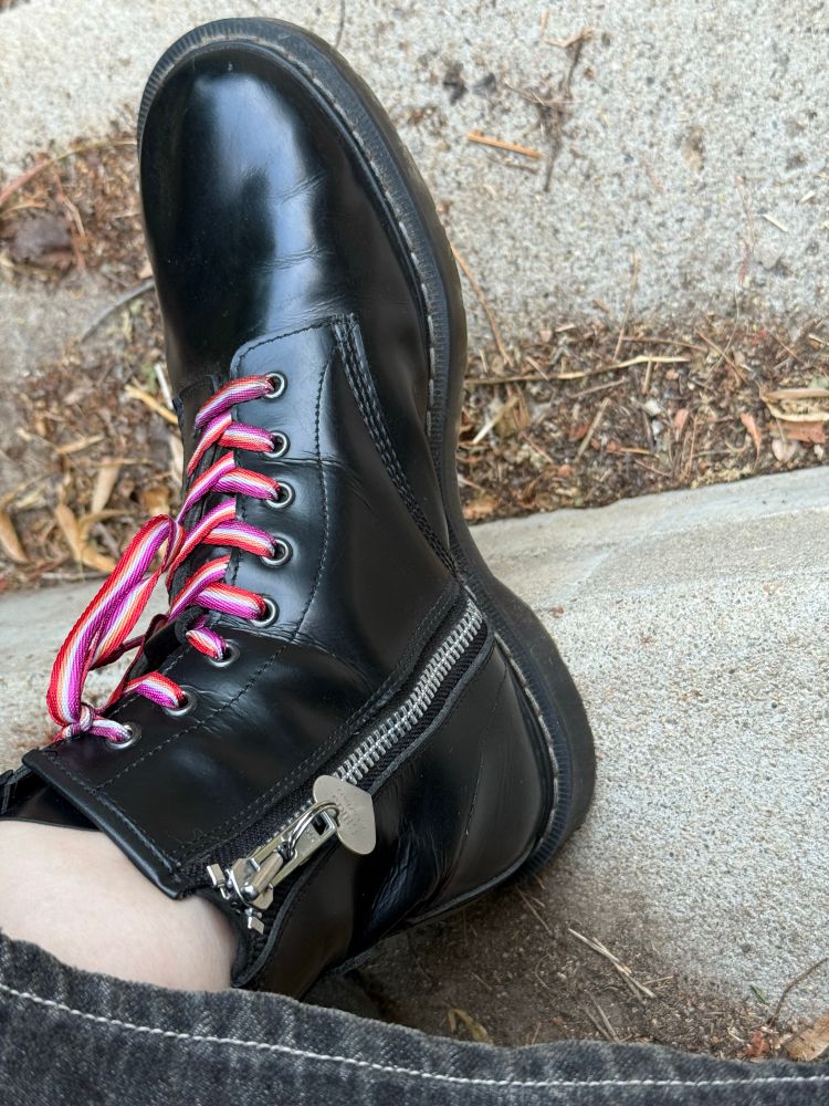 Picture of a black buffed side zip Solovair boot with lesbian flag colors and a heart shaped dog tag that reads Chris (she/her) attached to the pull.