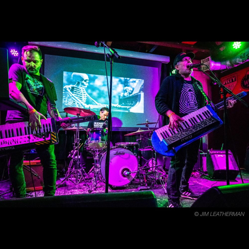 Live performance photo of Damage; image depicts Damage's three members, one on drums and two playing Moog Liberation synthesizers. Photo by Jim Leatherman.