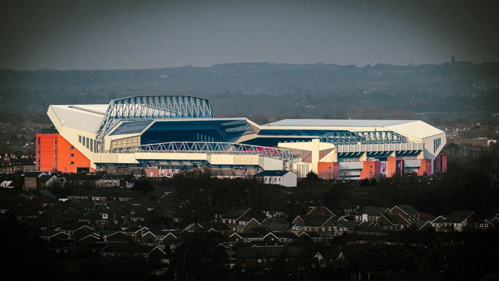 An zoomed image of Liverpool's football stadium seen from an elevated viewpoint over 2 miles away (~3.8 km). The image has been heavily edited to darken the foreground and background. The stadium has been selectively lightened to make it stand out.