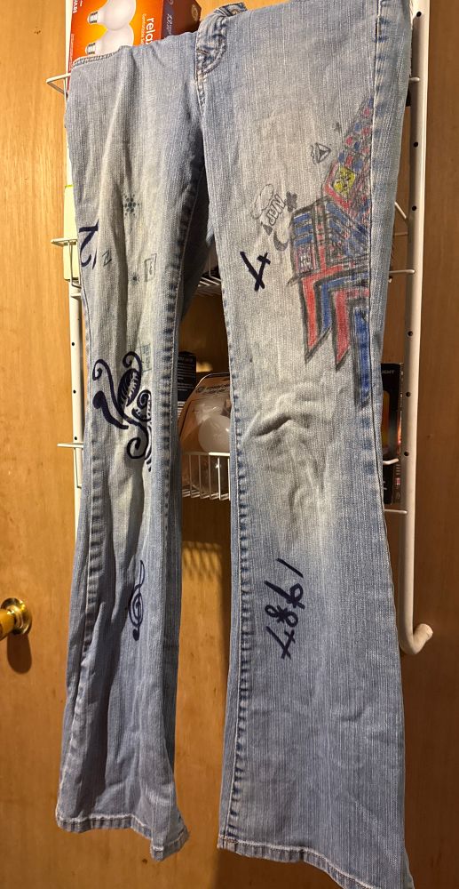 A pair of blue jeans with a flare cut and faded knees. The jeans are decorated with drawings and doodles in sharpie. Some of the doodles include the numbers “7,” “12,” and “1987;” as well as a treble clef; the word “NAP!” In bubble letters over a cloud and a large geometric red and blue pattern in the left thigh. 
