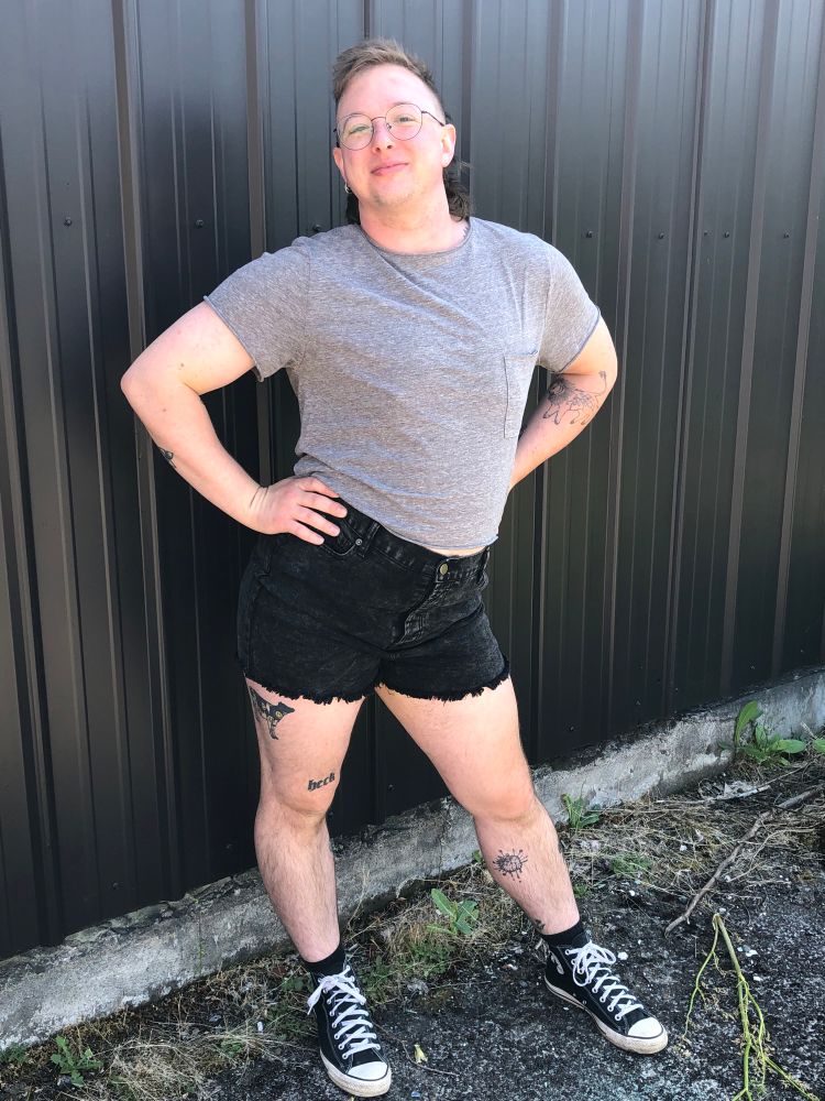 Lennox stands in front of a black corrugated metal wall with hands on their hips and a cute smirk on their face. They have a freshly trimmed light brown mullet hairstyle and are wearing wire-rimmed glasses, a gray crop tee, black cutoff shorts, and black Converse sneakers. 