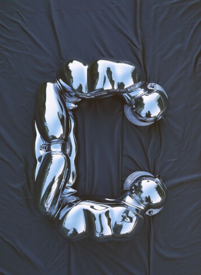  A silver, robotic-looking humanoid figure contorted into the shape of the letter "C," lying on a black fabric backdrop.