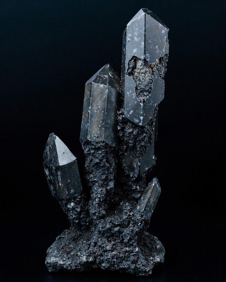 A vertical, dark quartz cluster with multiple pointed crystals protruding from a rough, rocky base, set against a black background.