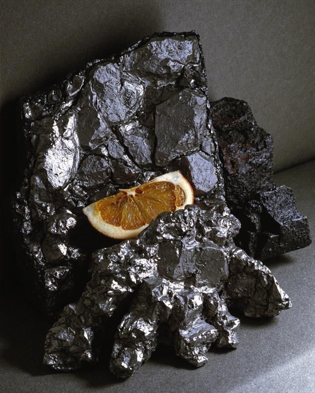 A still life composition of dark, metallic, rock-like formations with a dried orange slice resting among them. The textures and contrast evoke an abstract vision of the planet Mercury—harsh, reflective, and touched by fleeting warmth.