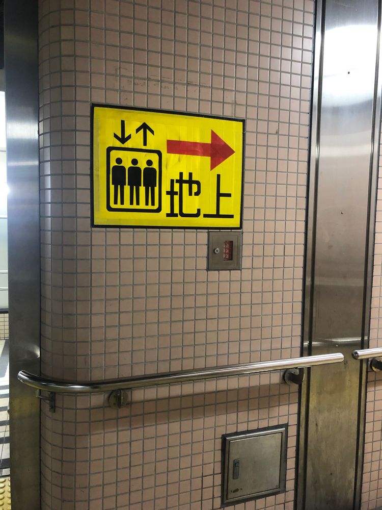 地下鉄構内の壁面の画像。くすんだピンクの正方形のタイルの壁面に、エレベーターを示す黄色い貼り紙と、手摺がある。