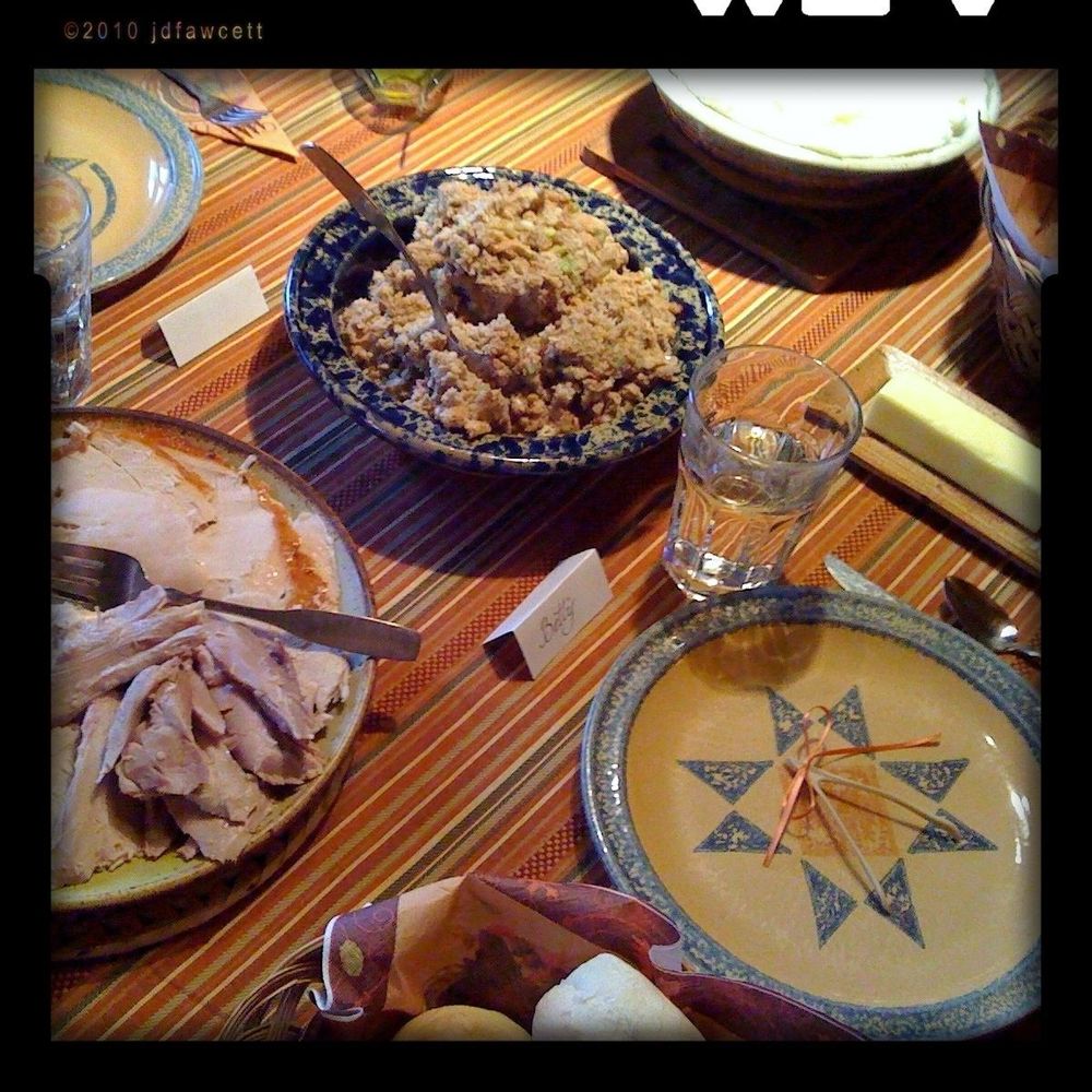A warmly lit table set for a shared meal features a homey, rustic atmosphere. The table is covered with a striped tablecloth in warm tones of red, orange, and gold. At the center sits a large, blue-patterned ceramic bowl filled with stuffing or dressing — moist, crumbly, and speckled with bits of celery or herbs. A metal serving spoon rests inside. To the left is a ceramic platter holding slices of carved turkey breast, still glistening with juices, with a fork and knife for serving. In front of the plate of stuffing is a small folded place card labeled “Betty” in handwritten script, suggesting this is part of a family gathering or holiday meal. A clear glass of water and a butter dish with a stick of butter on a wooden tray sit nearby. An empty plate with a star-like geometric pattern in blue sits at the bottom right, with the wishbone from a turkey arranged on top. A basket with dinner rolls or buns peeks in at the bottom of the image, lined with a festive napkin. Other ceramic dishes with indistinct contents are partially visible, and the warm lighting evokes an inviting, intimate setting typical of a Thanksgiving or similar holiday meal. 
