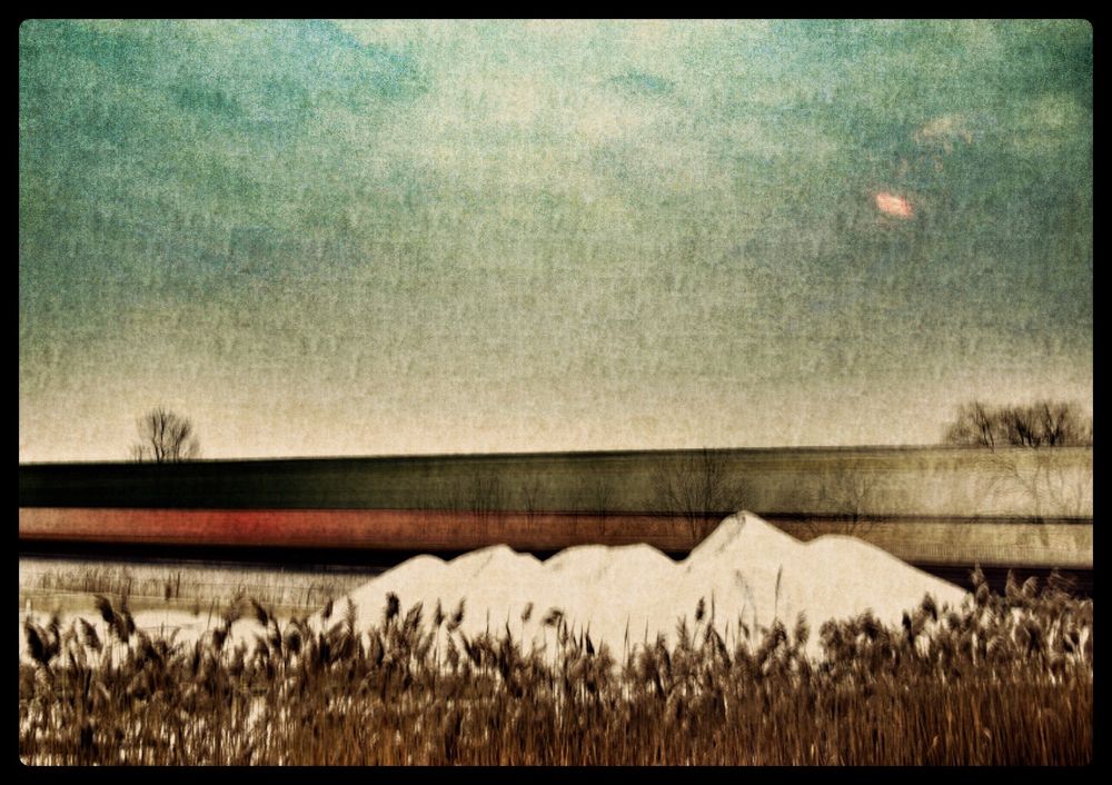 A surreal long exposure photograph captures a freight train passing through a barren landscape at dusk. In the foreground, tall grasses and reeds sway gently, their forms blurred and softened by the extended exposure. Behind them, a pale mound of snow or salt rises like a spectral hill, its texture glowing faintly under the ambient light. The train itself is rendered as a horizontal band of reddish motion, stretched across the middle of the frame like a glowing ribbon—its cars indistinct, transformed into a painterly smear of color and energy. Sparse, leafless trees stand scattered across the terrain, their silhouettes stark and skeletal against the muted backdrop. The sky above is a gradient of cool blues and greens, with a small, pinkish orb—possibly the moon—hovering near the horizon. A faint horizontal line cuts across the orb, suggesting a digital glitch or atmospheric distortion. The overall composition blends realism with abstraction, evoking a sense of quiet motion, winter solitude, and dreamlike passage.