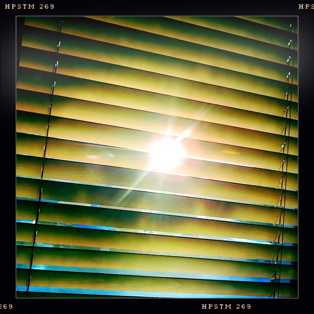 Close-up view of horizontal window blinds with sunlight streaming through the slats, creating a radiant burst of light and shadow interplay. The blinds are slightly tilted, allowing golden beams of sunlight to pierce through the gaps and illuminate the interior space with a warm, glowing intensity. The sun itself is visible through the slats, producing a strong lens flare effect that adds a sense of brilliance and optical drama. Light spills across the blinds in crisp, geometric bands, casting rhythmic shadows that echo the structure’s linear form. The outside world is partially visible through the openings, hinting at a clear sky and bright day beyond. The image is framed with a black border labeled “HPSTM 269,” suggesting a stylized or filtered capture from a retro-inspired camera app. The overall mood is luminous and uplifting, with the sunlight dominating the composition and evoking themes of clarity, energy, and awakening.