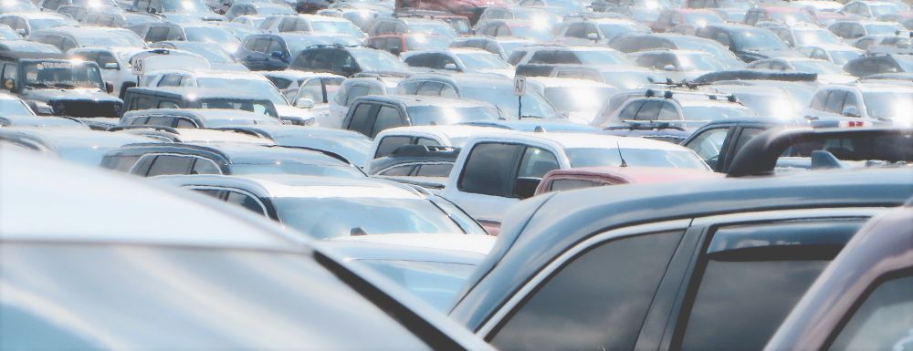 A vast parking lot crammed full of vehicles stretches into the distance, creating a dense sea of car roofs, windshields, and side windows that reflect bright sunlight. The cars, trucks, and SUVs are packed so tightly that individual lanes are barely distinguishable, making the scene feel almost chaotic and overwhelming. The shiny reflections from the glass surfaces give the impression of countless shimmering points of light, adding a slightly surreal or dreamlike quality to the image. Parking markers on poles, labeled “A7” and “A8,” rise above the mass of vehicles to designate sections, hinting at the organized structure beneath the apparent congestion. The muted colors—grays, whites, blues, and silvers—dominate the palette, with only a few darker or red vehicles punctuating the metallic monotony. The perspective compresses the scene so that the cars appear stacked and endless, evoking the feeling of vastness and the immense scale of modern car culture.