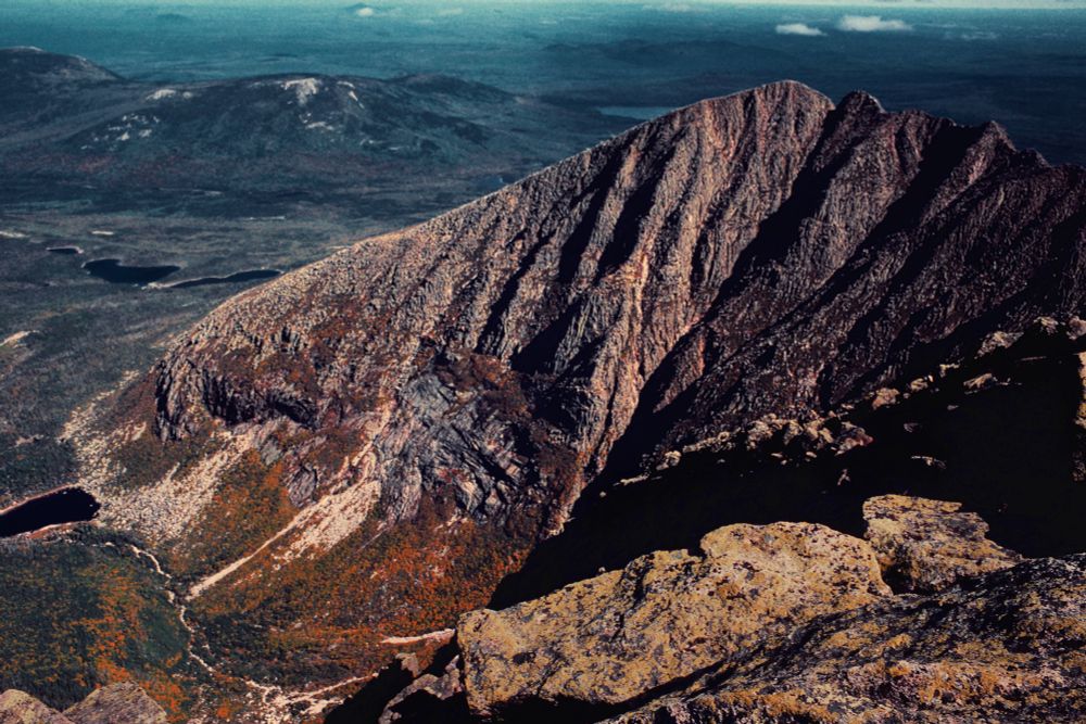 A panoramic view of the narrow Knife Edge Trail on Mount Katahdin, showcasing the rugged and dramatic landscape. The foreground features rocky terrain with large boulders, hinting at the challenging nature of the hike. The Knife Edge Trail, a thin ridge, extends along the mountain's summit, flanked by steep drop-offs on either side. In the background, the mountain's jagged peaks rise sharply, their surfaces marked by distinct geological layers and textures. The lower slopes are covered in a mix of green foliage and autumnal colors, suggesting a diverse ecosystem. In the distance, rolling hills and valleys stretch out under a clear blue sky, emphasizing the vastness of the natural environment. The overall scene conveys a sense of adventure and the beauty of the wilderness.