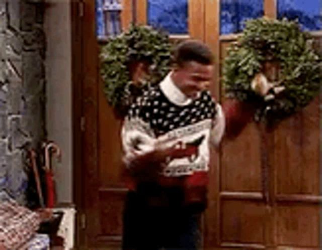 a man in an ugly christmas sweater is standing in front of a door with a wreath on it .