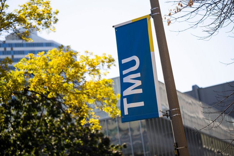 A close-up shot on a pole with a vertical flag indicating the logo of Toronto Metropolitan University with trees and a building in the background.