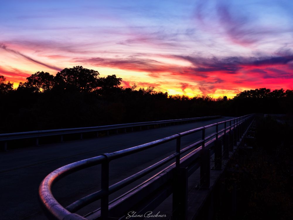 A bridge in the Government Canyon State Natural Area with a brilliant sunset sky overhead that is washing the bridge's siderails with colored light reflections.