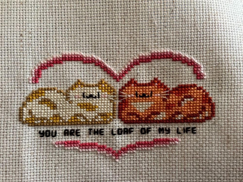A cross stitch of a yellow and white spotted cat and an orange tabby cat sitting in a pink heart. The words “you are the loaf of my life” are stitched below 