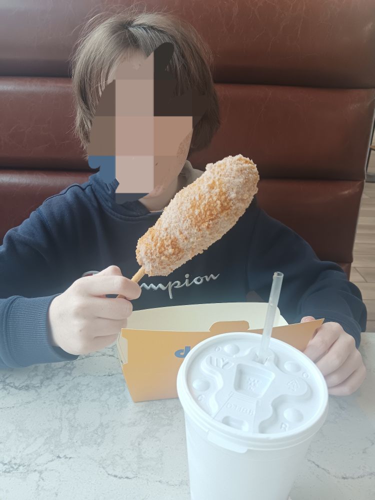 A boy in a blue hoodie holding a Korean corn dog filled with cheese and dusted with sugar.