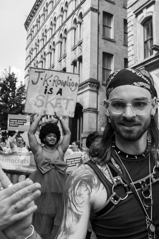 A Black and white photo of a Manchester street.
The masc person in the foreground is wearing a bandanna, vet and bondage straps, they are smiling and high-fiving a perosn out of frame. 
In the mid ground there is a person with a Afro, beard and a ball gown holding a sign that says ‘J K Rowling is a sket’ above their head. They look proud. 
In the background you can see the Liberal Democrat group with Trans Right signs and banners.