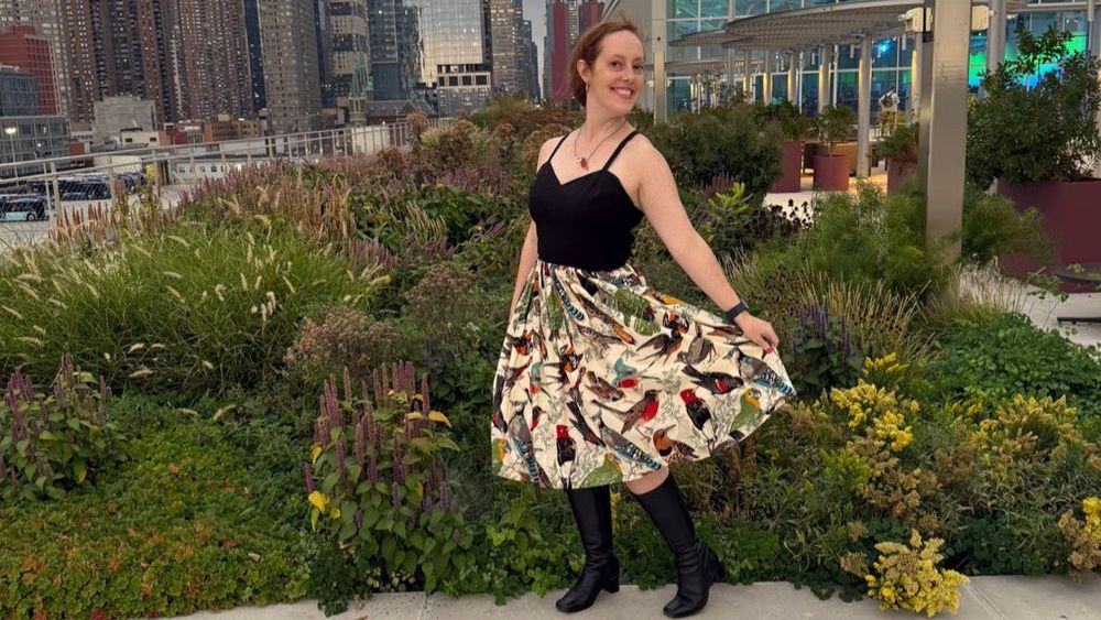 Robyn posing ostentatiously on the green roof at the Javits Center with NYC in the background wearing a dress with a black bodice and a skirt with a bunch of songbirds on the skirt and also very good boots 