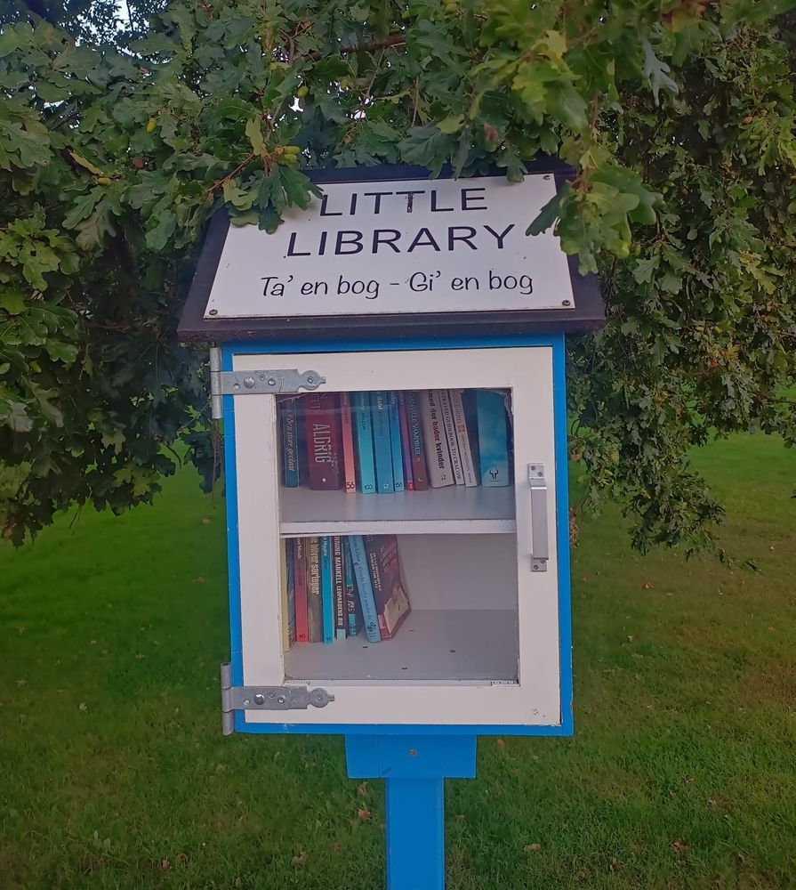 A blue-and-white wooden little library in green surroundings with a glass insert in the door. Inside around 20 books. A sign on the roof of the library box says "ta' en bog - gi' en bok" meaning "take a book, give a book"