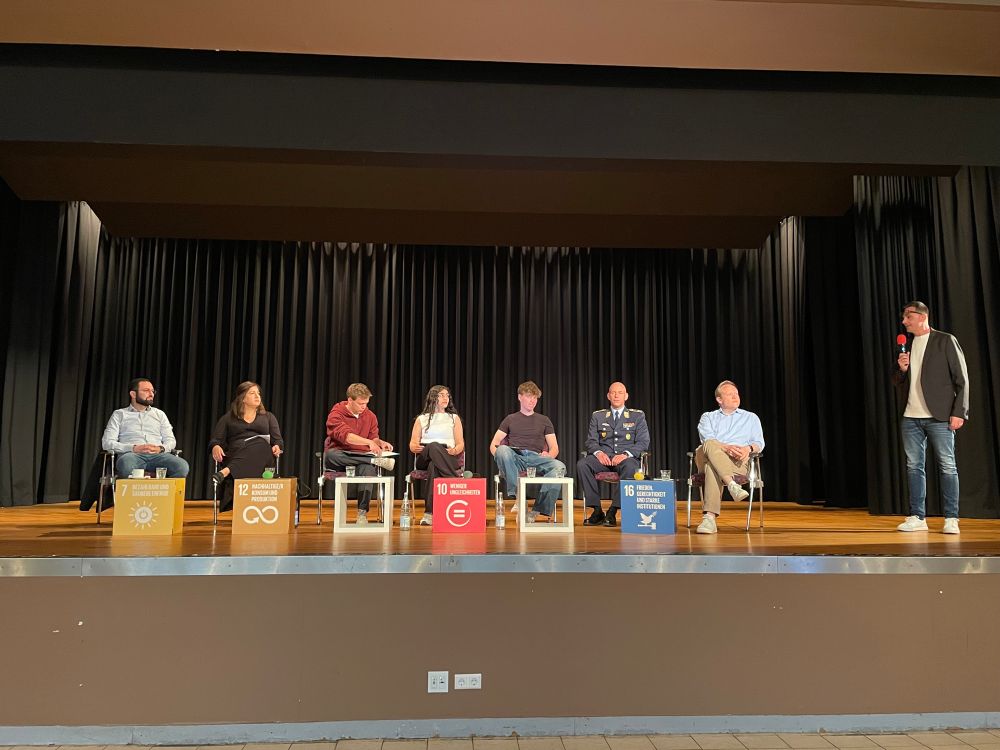 Bühne mit 7 Menschen auf Stühlen, eine Person steht und begrüßt die Gäste 