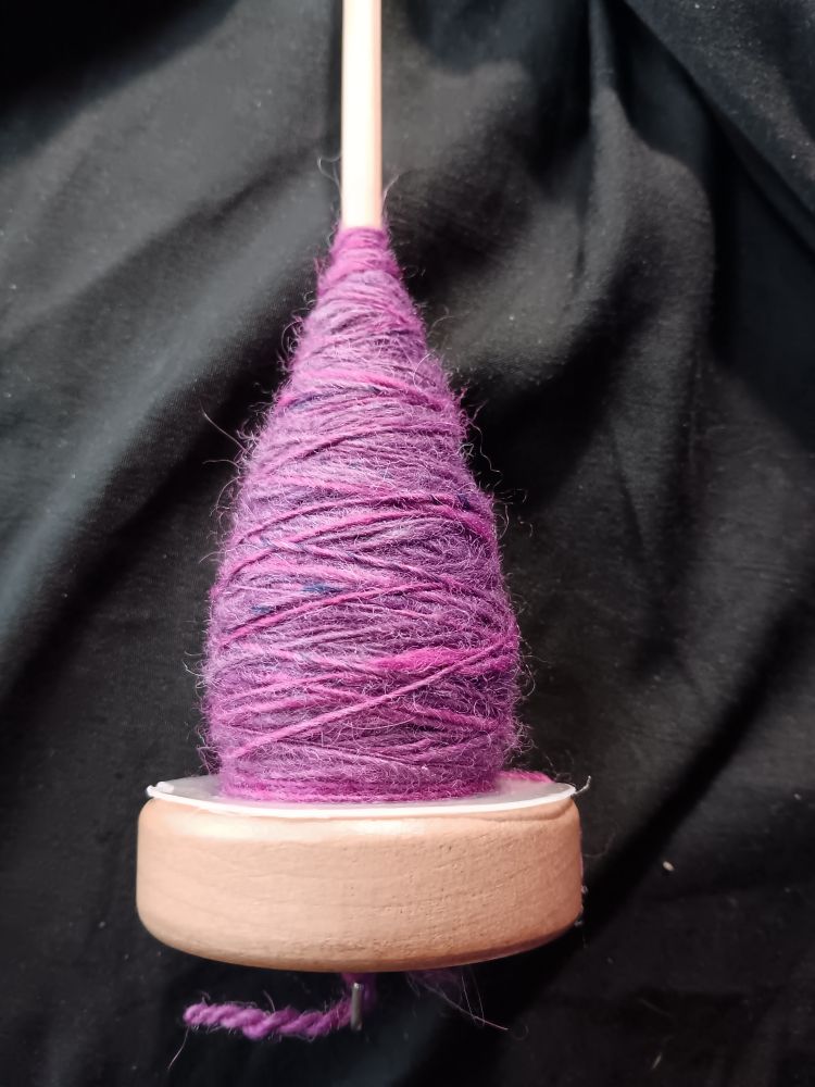 A drop spindle with a pear-shaped cop of yarn wrapped around the shaft. The yarn is a single-ply, and various shades of purple and pink. 

#yarn #handspunyarn #handspun #handspinning #fiberarts #handmade #spinnersofbluesky #spindlespinning #alpaca 