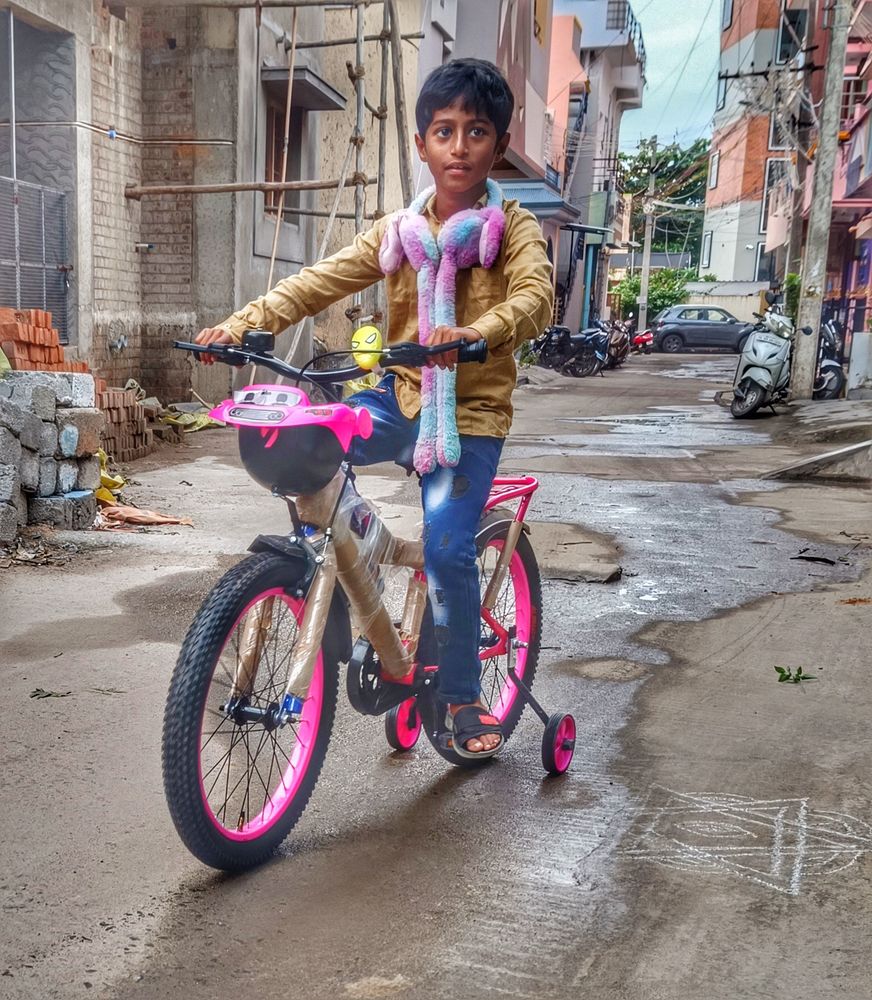 Saint Nicholas Day, 6 December.

A young boy sits on a pink and black bicycle with protective plastic wrap on the frame and training wheels attached to the rear axle.

He is wearing a gold-colored long-sleeve shirt, blue jeans, and a colourful plush scarf. 

The lane is in a residential area with buildings and scaffolding in the background. Ramana Nagar, Tiruvannamalai, Tamil Nadu, India.