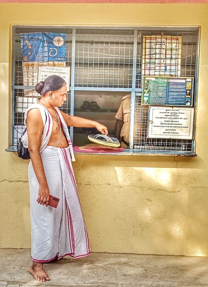 A young Indian Brahmin dressed in a white dhoti and angavastram (a single rectangular fabric piece used for religious or ceremonial purposes), his hair tied in a neat bun, a black handbag over his left shoulder and a cellphone in his right hand, and wearing a holy thread, a necklace and earrings, deposits his grey and yellow chappals (sandals) at a footwear stand. He carries a cellphone in his right hand.

The English text on one sign reads: "FOOTWEAR NOT ALLOWED", "PLEASE LEAVE IT IN THE FOOTWEAR STAND", and "KEEP SILENCE".

Sri Ramanasramam, Chengam Road, Tiruvannamalai, Tamil Nadu, India.