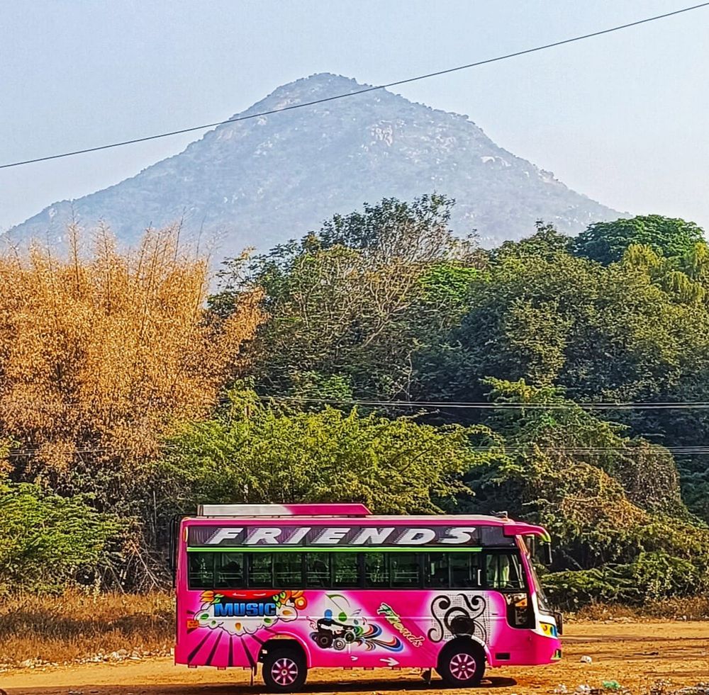 A pink, 40-seat private bus with a large 'FRIENDS' decal on the upper window sits parked on a clay plot of land with Arunachala looming in the background, separated only by a variety of trees and dense undergrowth.

The holy hill forms part of the Eastern Ghats and is 2,268 feet in height. Geologists estimate its age to be 2.6 billion years. The Skanda Purana tells of how Shiva appeared as a column of light to settle a dispute between Brahma and Vishnu about which of them was greater. After this had been resolved, Siva manifested in form for the benefit of humanity as the Arunachala Hill.