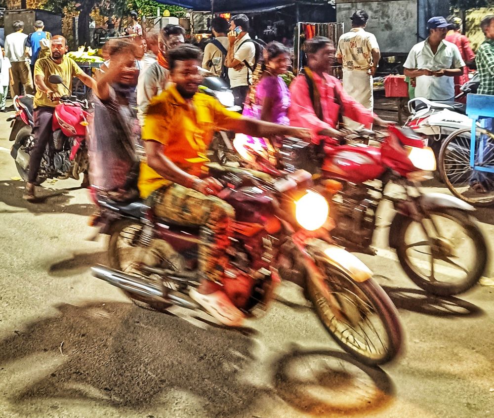 At a busy crossing point, a passenger on the back of a well-lit motorcycle waves in good humour. Of the three motorcycles in view, nobody is wearing a helmet. Behind them, street traders sell peanuts, coconut juice and vegetables to the passing trade. Tiruvannamalai, Tamil Nadu, India.