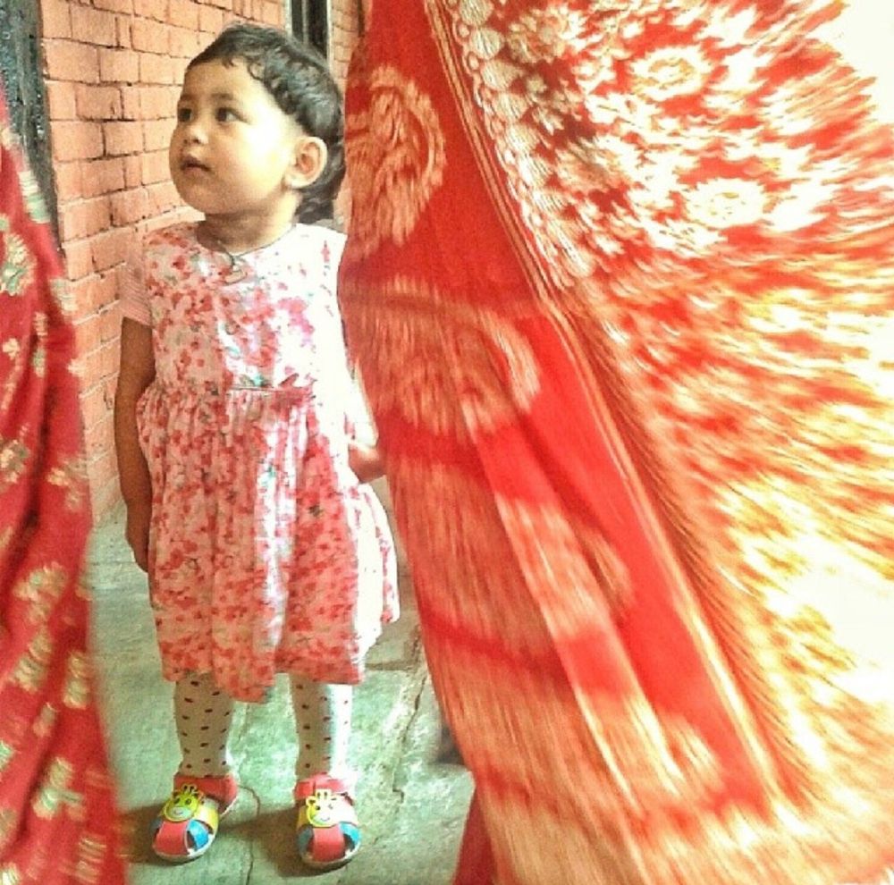 A short-haired child in a knee-length red floral dress, white stockings and multicoloured shoes gazes innocently between two women in red-patterned silk saris at a wedding in Kathmandu, Nepal.
