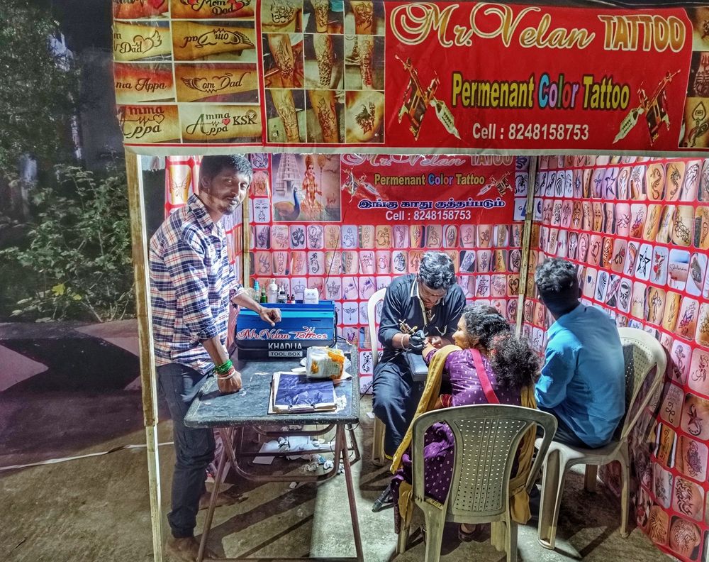 A temporary outdoor tattoo stall offers permanent colour tattoos and ear piercing services. 

The tattoo artist works on a woman's arm while a man, probably her husband, sits beside her looking at the display of tattoo designs on offer.

To the left, a third man stands and rests his arm on a blue metal box branded with 'Mr. Velan Tattoos' and 'Khadija' in decorative script. 