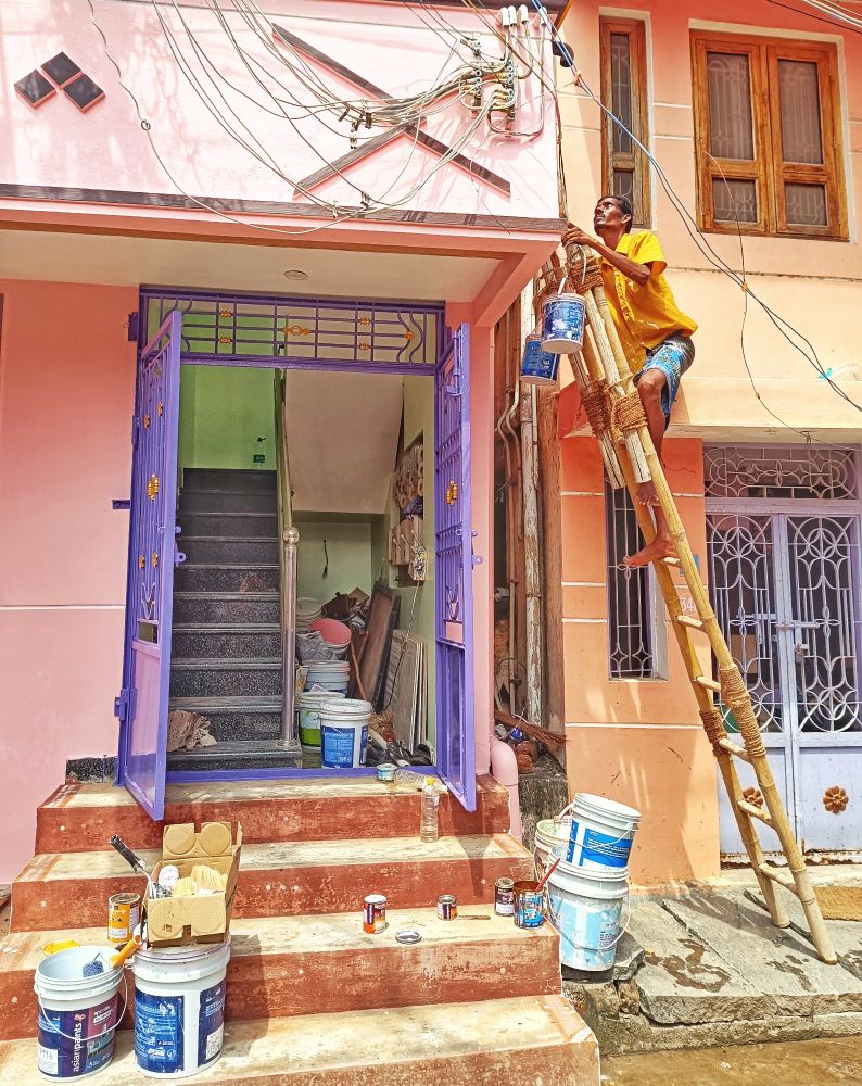 An Indian painter wearing blue shorts and a yellow tshirt perches on top of a bamboo ladder with buckets dangling from the top rung and leaning against an overhanging porch. The steps leading up to a staircase are covered with buckets, boxes and pots. Tiruvannamalai, Tamil Nadu, India.