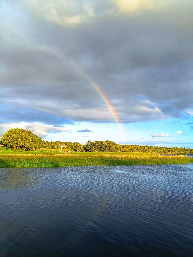 A half-arc of a rainbow passes through a mostly dark-grey sky with a streak of vivid bright blue. It is reflected on the surface of a passing river. In between, green rushes, lawns and trees dissect the image.