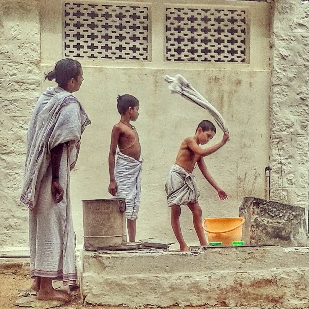 Two veshti-clad trainee brahmin priests look on as a third beats a cloth on the steps to clean it after soaking it in an orange plastic bucket. Sri Ramanasramam, Tiruvannamalai, Tamil Nadu, India.