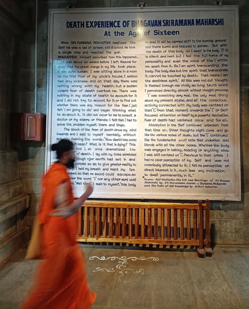 A man dressed completely in saffron clothing to symbolise his renunciation, detachment from worldly pleasures, and a life of penance and spirituality, walks past a spotlit hand-painted board with an account of the death experience as described by Bhagavan Sri Ramana Maharshi. 

When SRI RAMANA MAHARSHI realized the Self he was a lad of sixteen, still at school. He took a single step and reached the goal.

MAHARSHI himself described how this happened It was about six weeks before I left Madurai for good that the great change in my life took place. It was quite sudden. I was sitting alone in a room on the first floor of my uncle's house. I seldom had any sickness, and on that day there was nothing wrong with my health, but a sudden violent fear of death overtook me. There was nothing in my state of health to account for it and I did not try to account for it or to find out whether there was any reason for the fear. I just felt 'I am going to die' and began thinking what to do about it. It did not occur to me to consult a doctor or my elders or friends: I felt that I had to solve the problem myself. there and then.

The shock of the fear of death drove my mind Inwards and I said to myself mentally, without actually framing the words: 'Now death has come: what does it mean? What is it that is dying? This body dies: And I. at once dramatized the occurrence of death. I lay with my limbs stretched out stiff as though rigor mortis had set in and imitated a corpse so as to give greater reality to the enquiry. I held my breath and kept my lips tightly closed, so that no sound could escape so that neither the word 'I' nor any other word could be uttered. 

"Well then," I said to myself, "this body dead. It will be carried stiff to the burning ground and there burnt and reduced to ashes. But with the death of this body am I dead? Is the body 'I'? It is silent and inert but I feel the full force of my personality and even the voice of the I' within me apart from it. So I am spir…