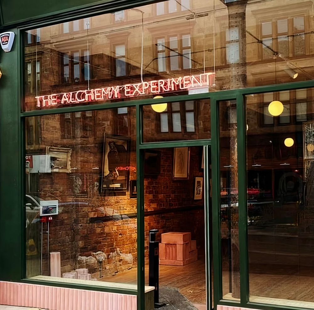 Exterior photo of The Alchemy Experiment shop from with green woodwork big, big windows and illuminated neon sign 