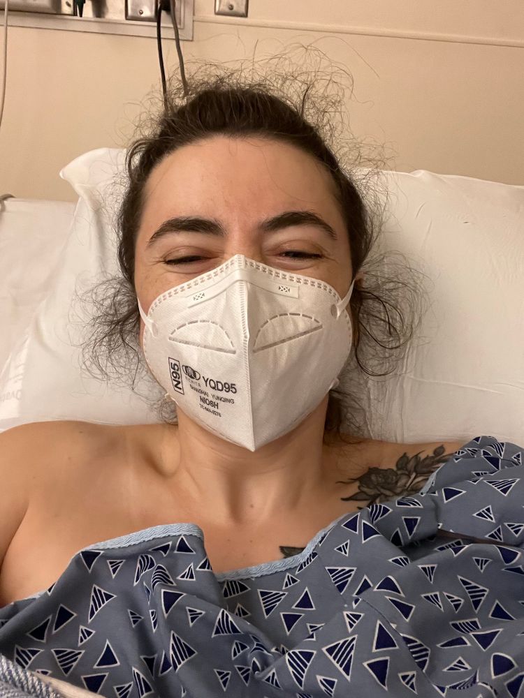 raea a white woman in a mask and a hospital bed and gown smiling widely