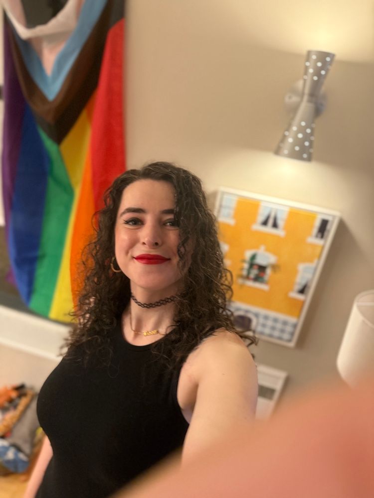 raea a curly haired brunette white woman in a sleeveless black dress and red lipstick smiling