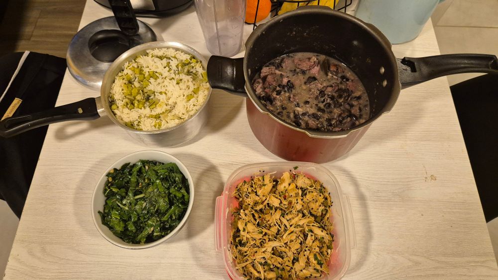 mesa quadrada e branca com comida em cima: no canto superior esquerdo, uma panela de alumínio com arroz branco e talos de couve; no canto superior direito, uma panela de pressão aberta com feijoada; no canto inferior esquerdo, um bowl de cerâmica com couve refogada; no canto inferior direito, um pote quadrado com peito de frango cozido, desfiado e temperado