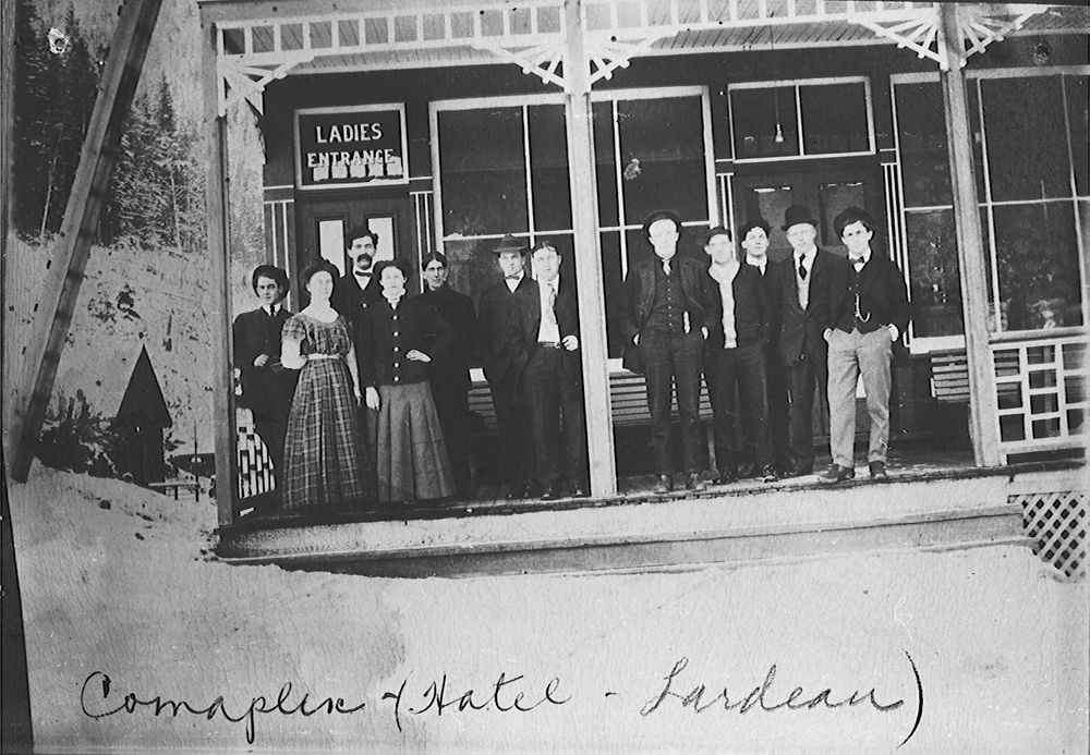 Photo of a group of men and two women standing outside the Landeau Hotel in Comaplix, BC around 1914. The hotel has a Ladies Entrance and is a simple building. Freda Evans is standing in the front row and wearing a long dress ran the hostel with her husband Russell. She was travelling on the Empress of Ireland, but did not survive the sinking.