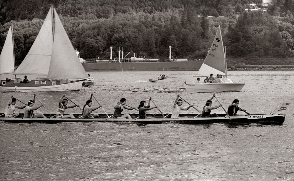 Nine Indigenous women compete in a traditional dug-out canoe named the St. Michael in the annual races at Cates Park in the mid-1970s. Two sail boats are alongside them and there is a ship in the background