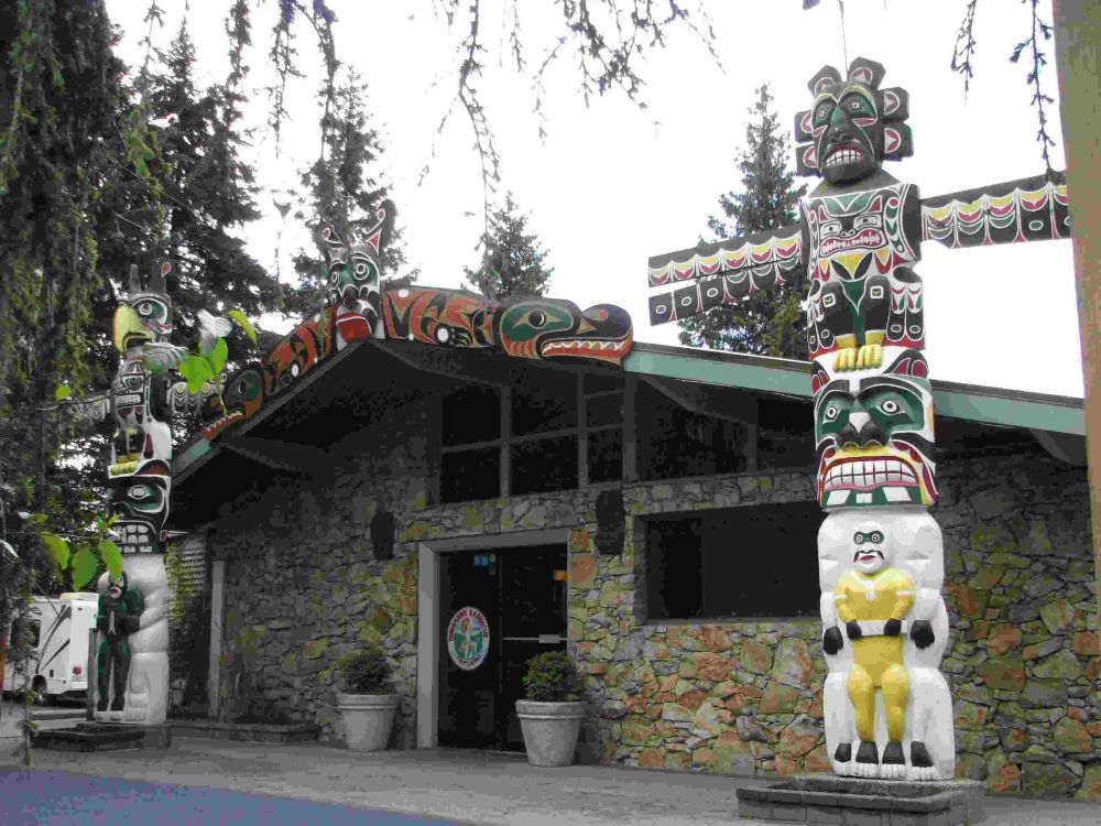 Photo shows a one storey building with stone on the outside, flanked by two huge totem poles carved in the 1930s
