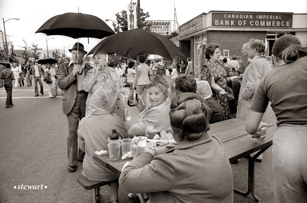 A man in a suit and hat is holding an umbrella, a woman is sitting and holding an umbrella while and a group of men and women are standing and sitting along West Broadway eating food at one of the first Greek Days in Kitsilano in the mid 1970s. 