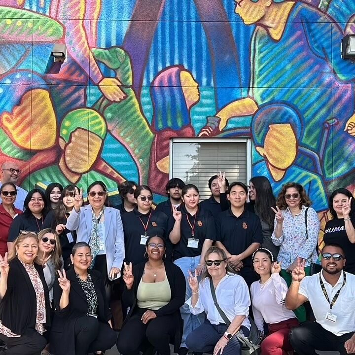 A group of youth and adults holding up peace signs and smiling while standing in front of a mural.