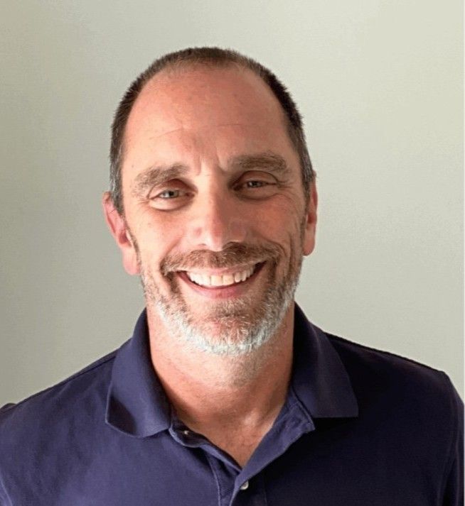 Portrait of a smiling individual with a trimmed beard, wearing a navy blue shirt, standing against a light gray background.