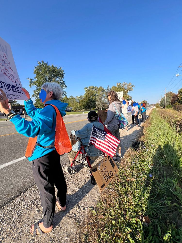 Protesters lined up along roadside, one with an American flag. Signs about not allowing constitutional sheriffs being held.
