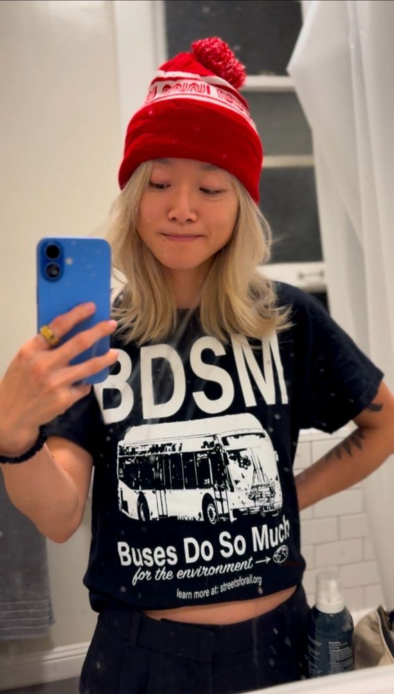 Woman wearing a MUNI beanie and a shirt that says “BDSM: buses do so much”
