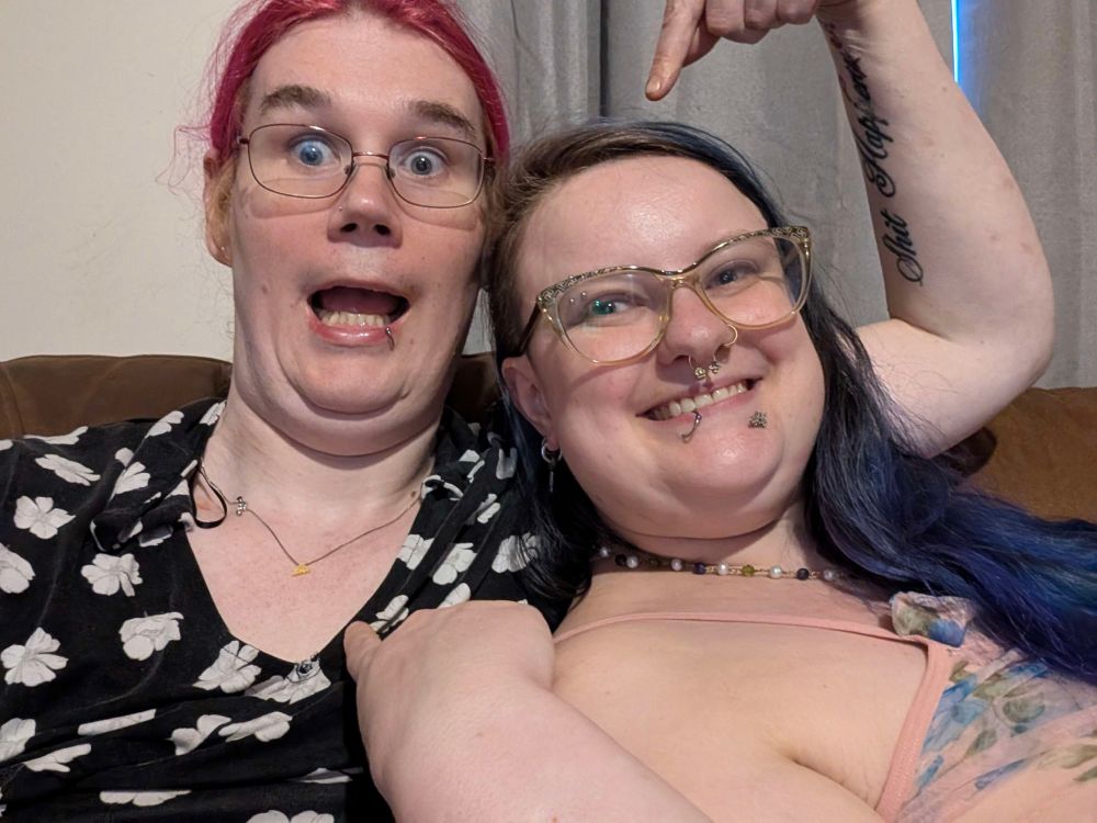 lexi and sinéad are sitting next to each other on couch, shown from front chest-up. sinéad has pink and blue floral nightie and genderqueer beaded necklace. lexi is wearing same dress as previous, along with a silver chain with delicate yellow pendant, which sinéad is pointing at while lexi points at sinead’s head with arm with “shit happen[ed]” tattoo. lexi has eyes open super wide and an open mouth silly-face beam and sinéad is grinning very gayly (pun intended). 