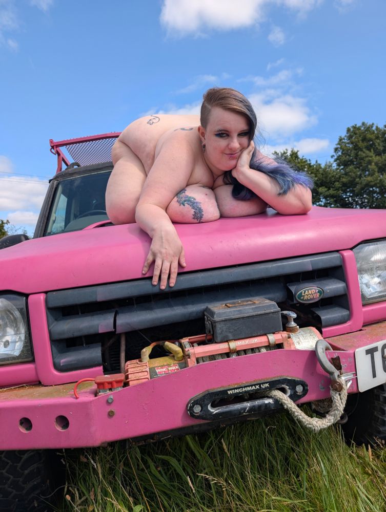 slight low angle looks up at a blue land rover that’s mostly been painted bright as fuck pink, with a winch on the front and pink roof rack on top. the vehicle is in a grassy field with blue sky above and trees in background. on the hood of the land rover is sinéad, on faer front facing the camera, ass up and tits supporting chest and shoulders as fae drapes faerself over the pink car. faer head is on left hand, elbow to pink surface, with right hand dangling casually over the grill. faer boob tattoo is visible and fae is smirking. 